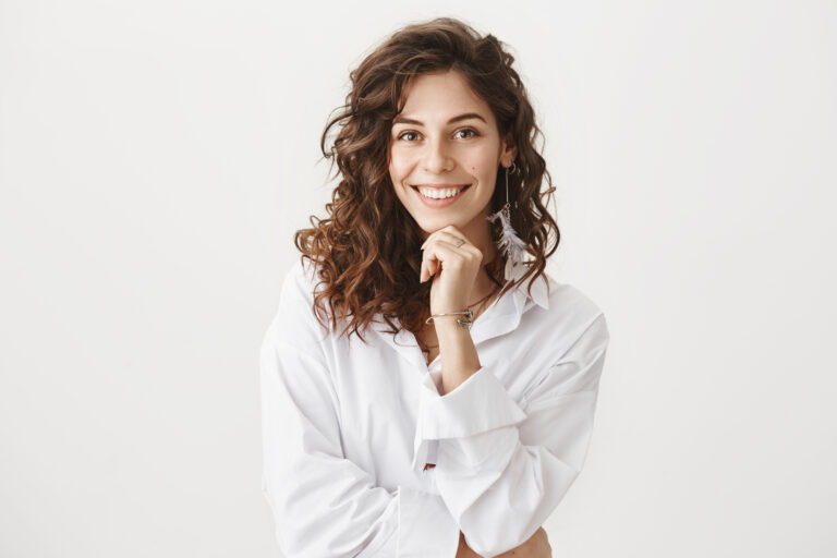 Cute happy european woman with curly hair in trendy white blouse, smiling broadly, leaning chin on hand, listening with interest to friend story, standing amused and joyful over gray wall